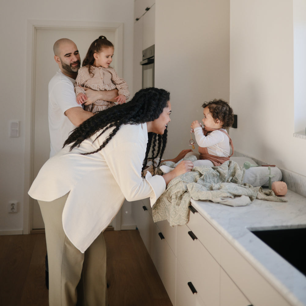 Lifestyle image of a young family of four gathered around a kitchen counter with a young boy sitting on a changing pad  with his mother leaning down and smiling at him. A Cambridge Blue Star Training Toothbrush and Blush Dice Press toy are laid by the baby. 