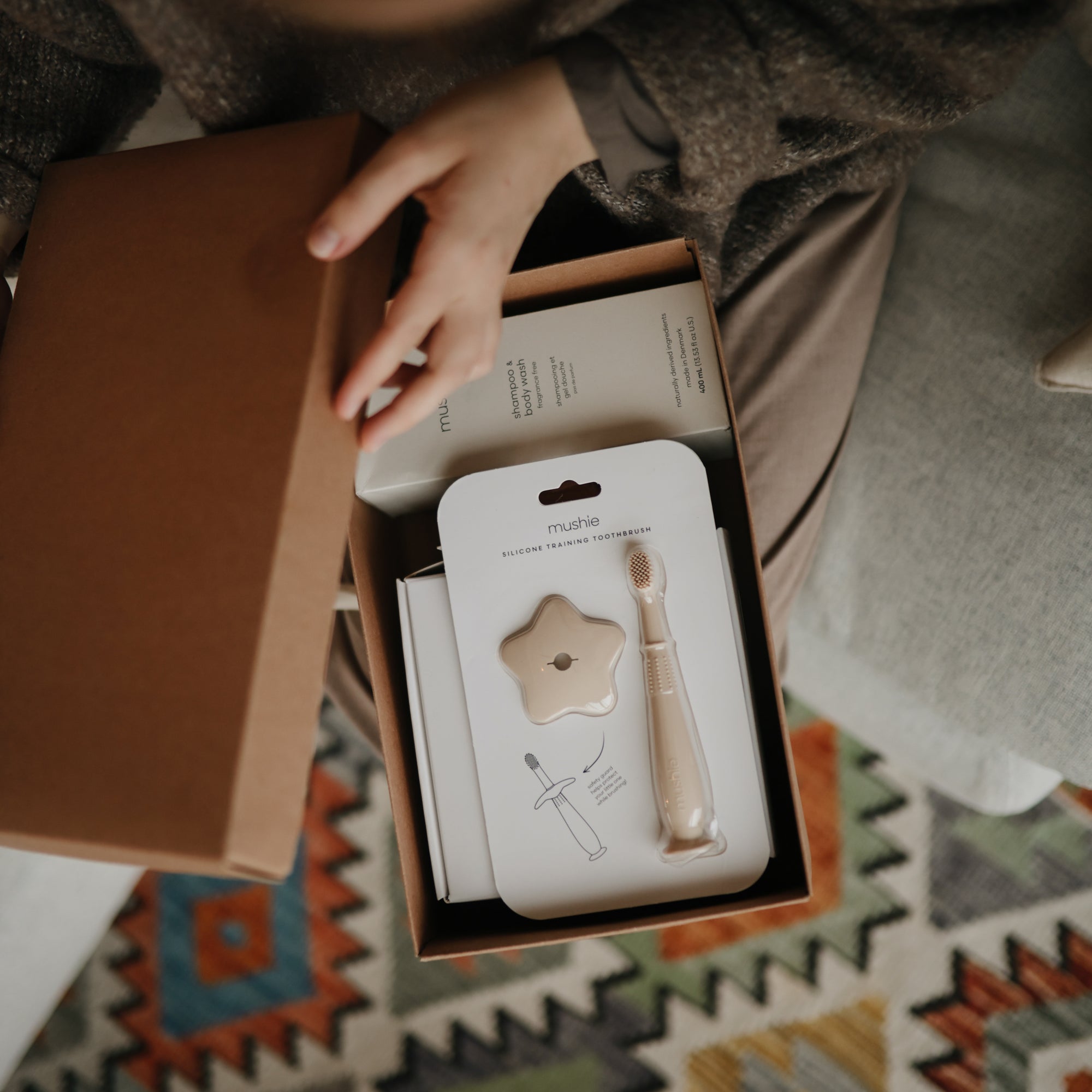 Lifestyle image looking into a Mushie gift box in a person&#39;s lap as they lift the lid off the box, revealing a Shifting Sand Star Training Toothbrush and Baby Shampoo in full packaging. 